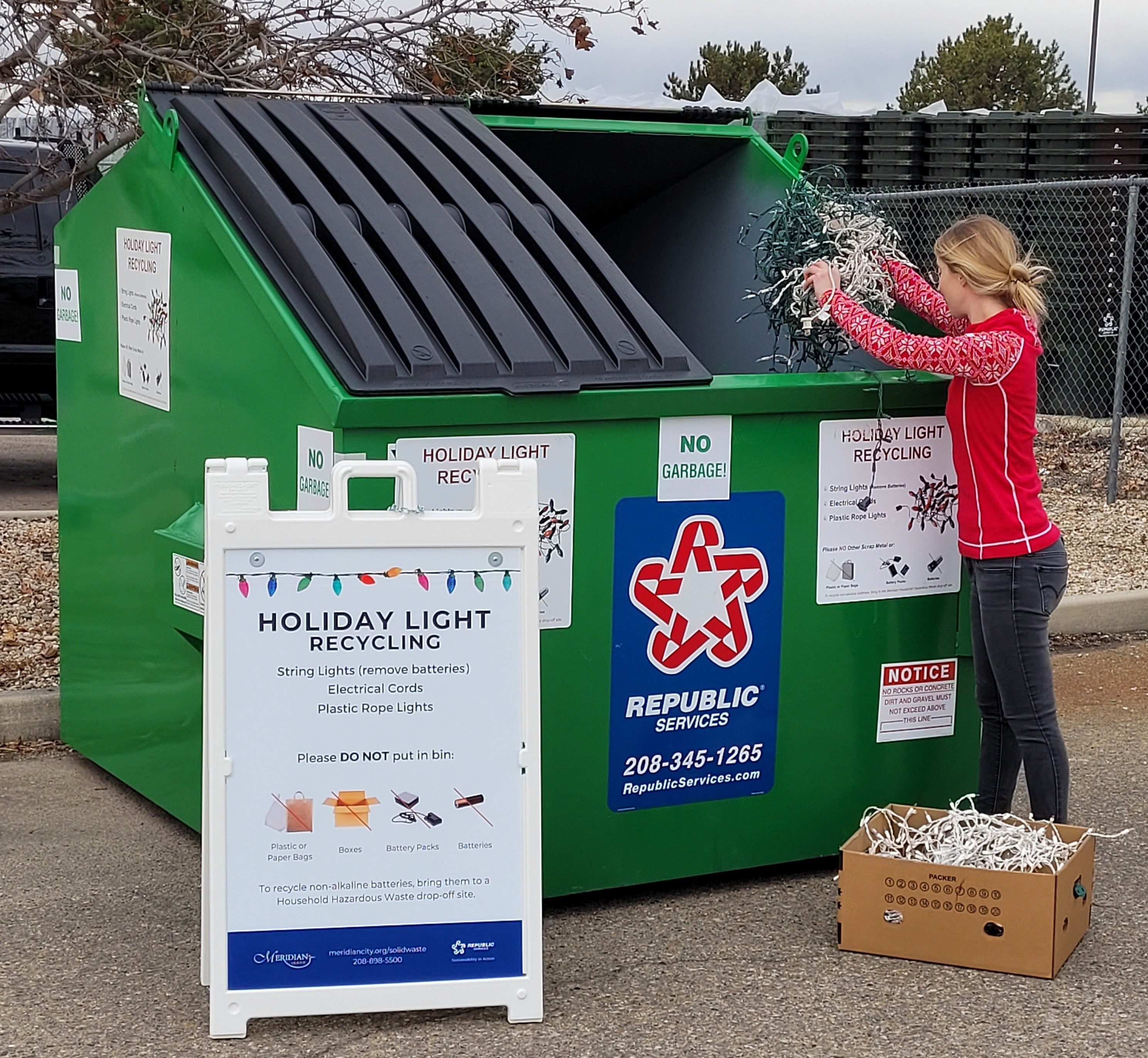 Person placing string holiday lights in a large, green Holiday Light Recycling bin